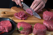 Person cutting raw beef on a wooden board with a knife, surrounded by more beef and vegetables.