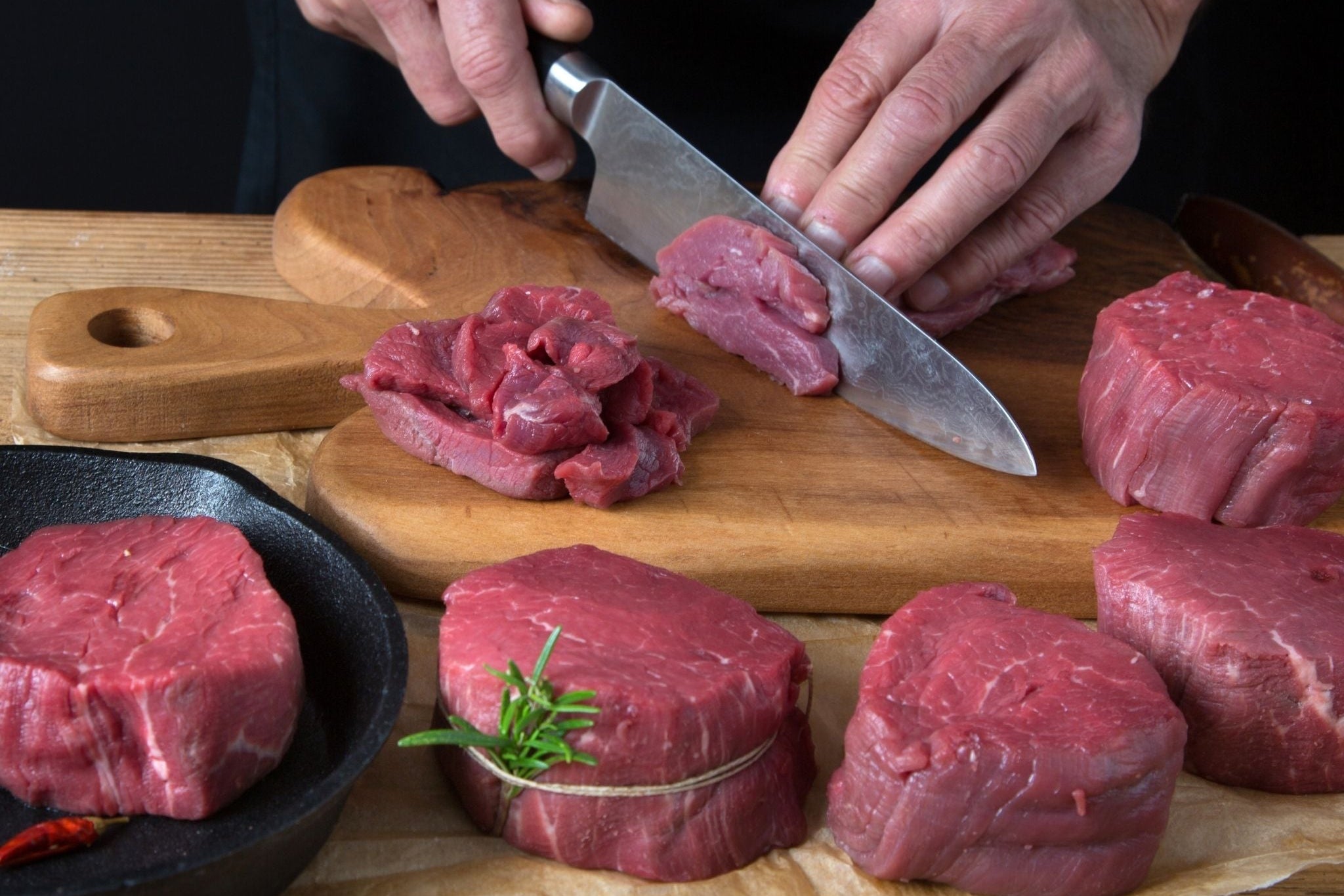 Person cutting raw beef on a wooden board with a knife, surrounded by more beef and vegetables.